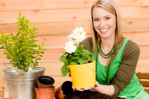 The image depicts a smiling woman with blonde hair in an outdoor garden setting, holding a small yellow flower pot with white flowering plants, possibly potted geraniums or similar flowering shrubs. She is dressed in a green top with a matching green beaded necklace and a brown cardigan. In front of her on a wooden surface are several empty plant pots, including a small black one and an orange terracotta one. To her left, there is a larger, metallic garden pot containing a lush, green shrub with dense foliage. The background features a wooden fence, indicating a neatly maintained garden area with natural lighting suggesting a bright, sunny day. The setting emphasizes a well-organized outdoor space suitable for gardening activities, such as planting, potting, or general garden maintenance, aligning well with professional gardening and landscaping services offered by Hedge Trimming Eltham in the Eltham area.