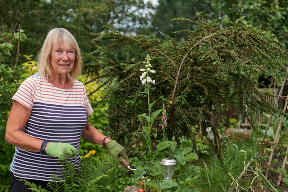 An elderly woman with shoulder-length blonde hair, wearing a short-sleeved striped shirt in white, navy, and red, and green gardening gloves, is standing in a lush garden. She is holding garden shears