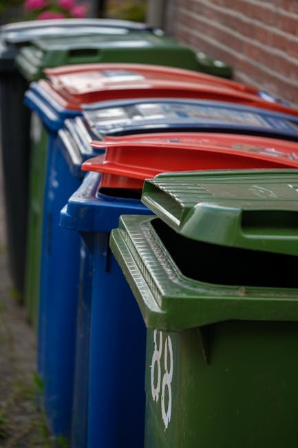 A row of large wheelie bins lined up along a garden pathway, including blue, green, red, and grey containers, with some lids open revealing garden waste such as leaves, grass clippings, and small bran